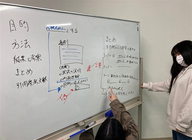 ホワイトボードを指さしながら、研究の相談をする2名の学生